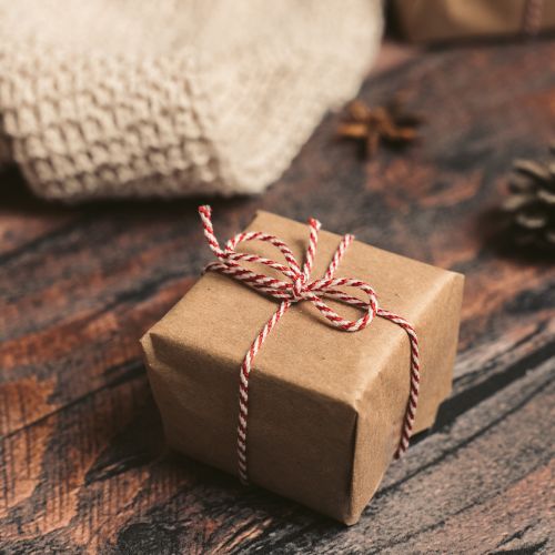 A small box wrapped in kraft paper and tied with red and white twine in a neat bow, resting on a wooden table with a soft blanket draped to the side.