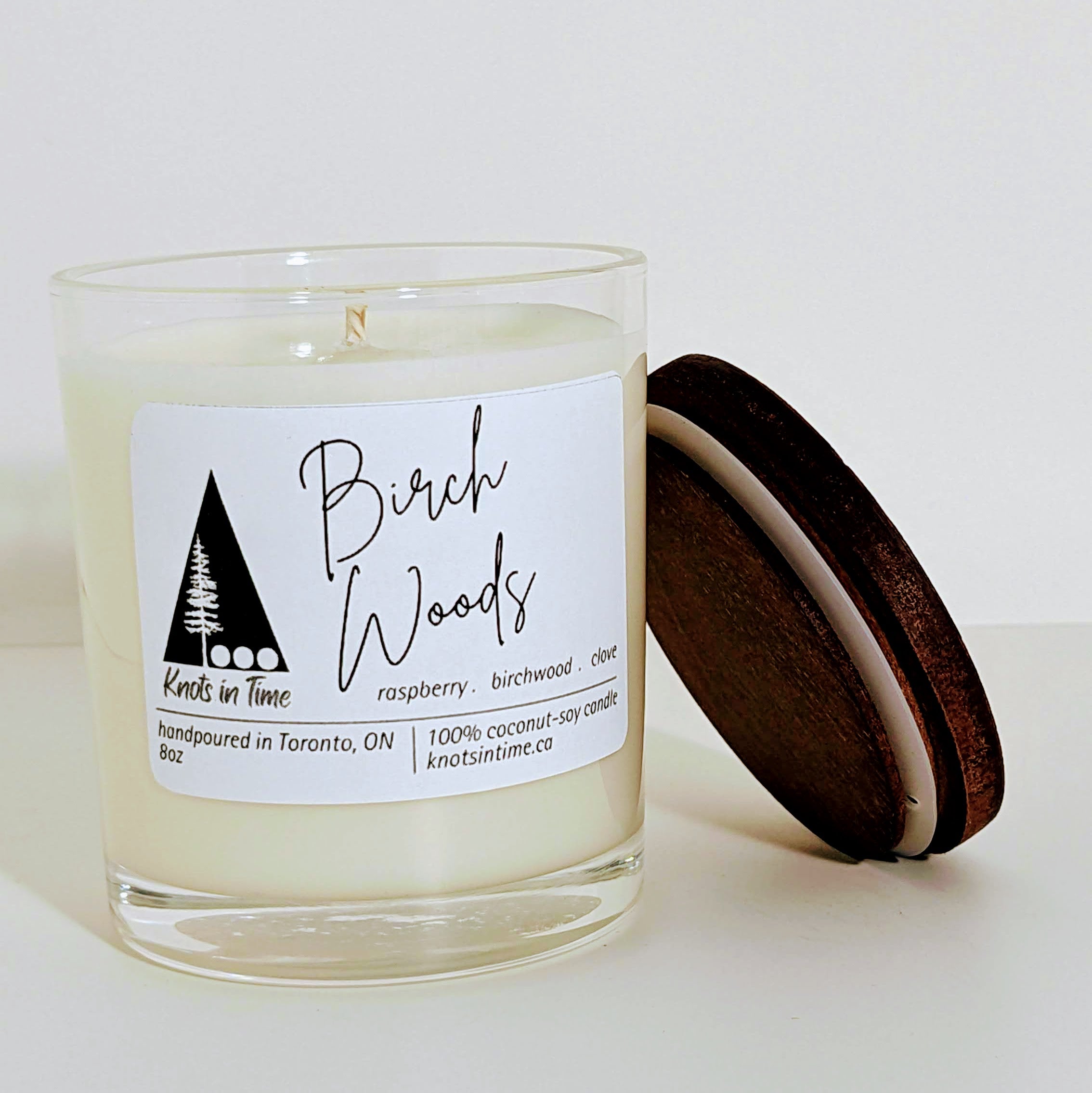 An unlit 8oz clear glass candle with a white label featuring a company logo and scent name, with a wooden lid leaning against its side. The candle is set against a plain white background.