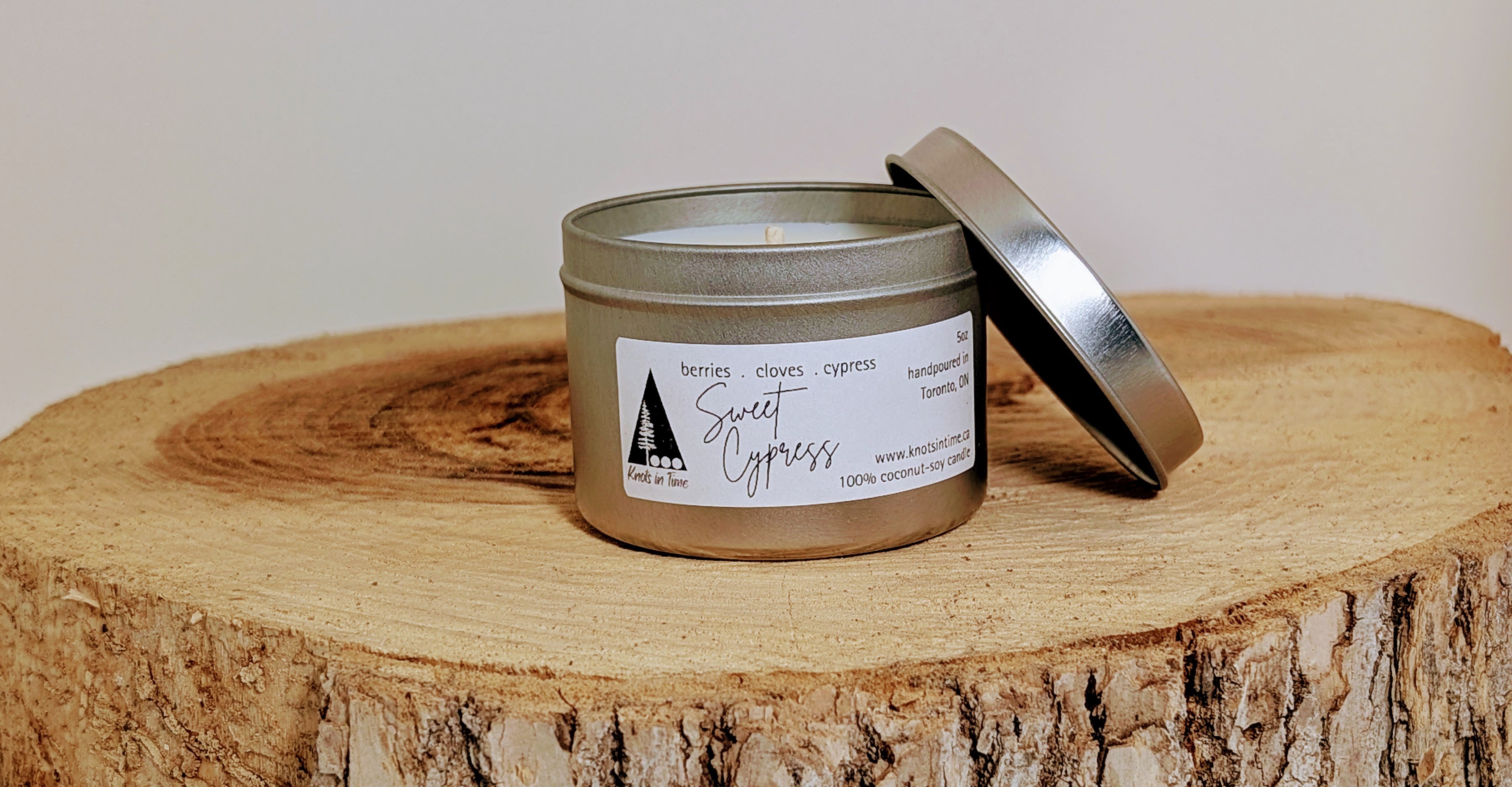 A silver tin candle sits on a round wood slice, with the lid propped against the side of the tin. The natural wood grain contrasts with the sleek metal finish, creating a rustic and cozy feel.