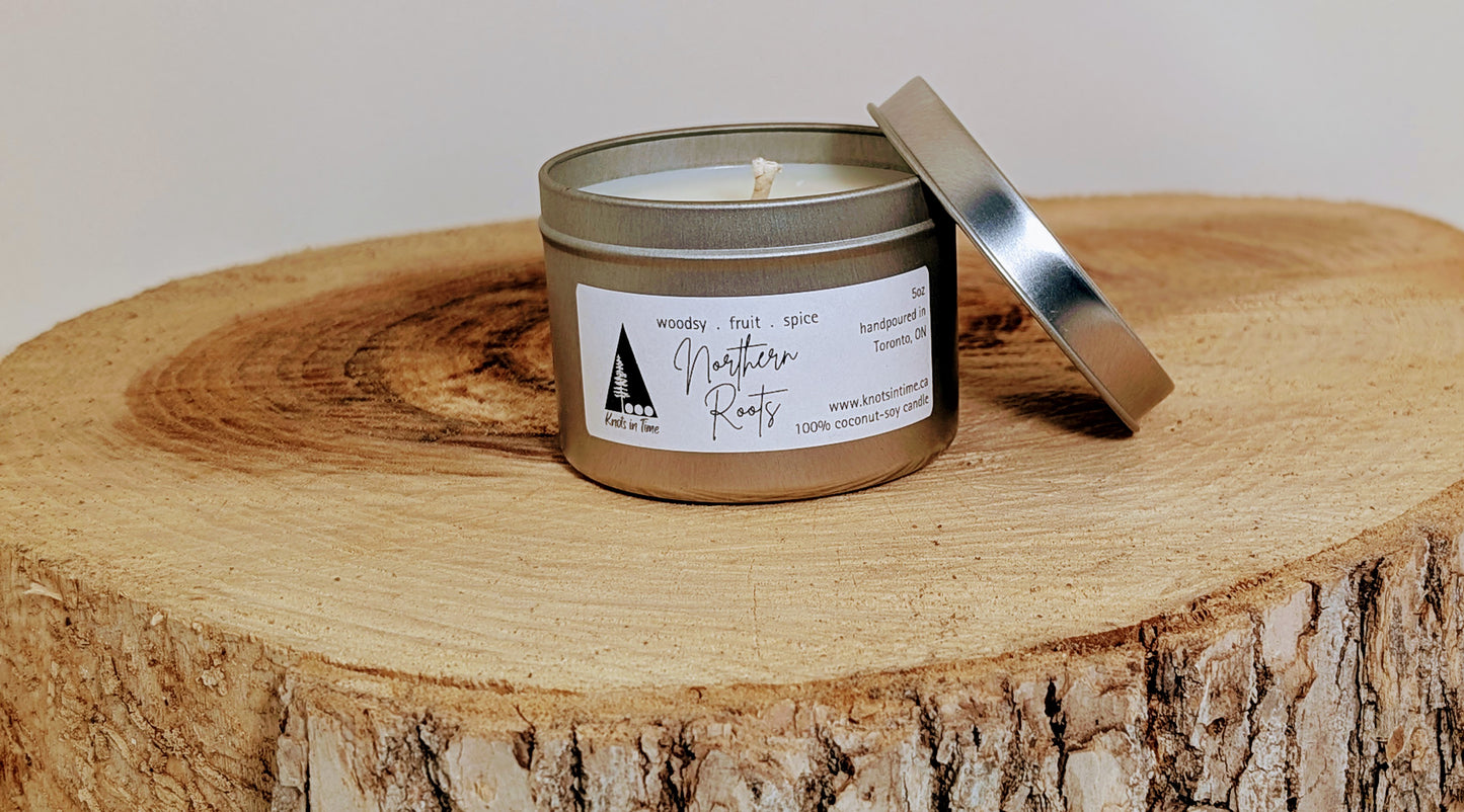 A silver tin candle sits on a round wood slice, with the lid propped against the side of the tin. The natural wood grain contrasts with the sleek metal finish, creating a rustic and cozy feel.