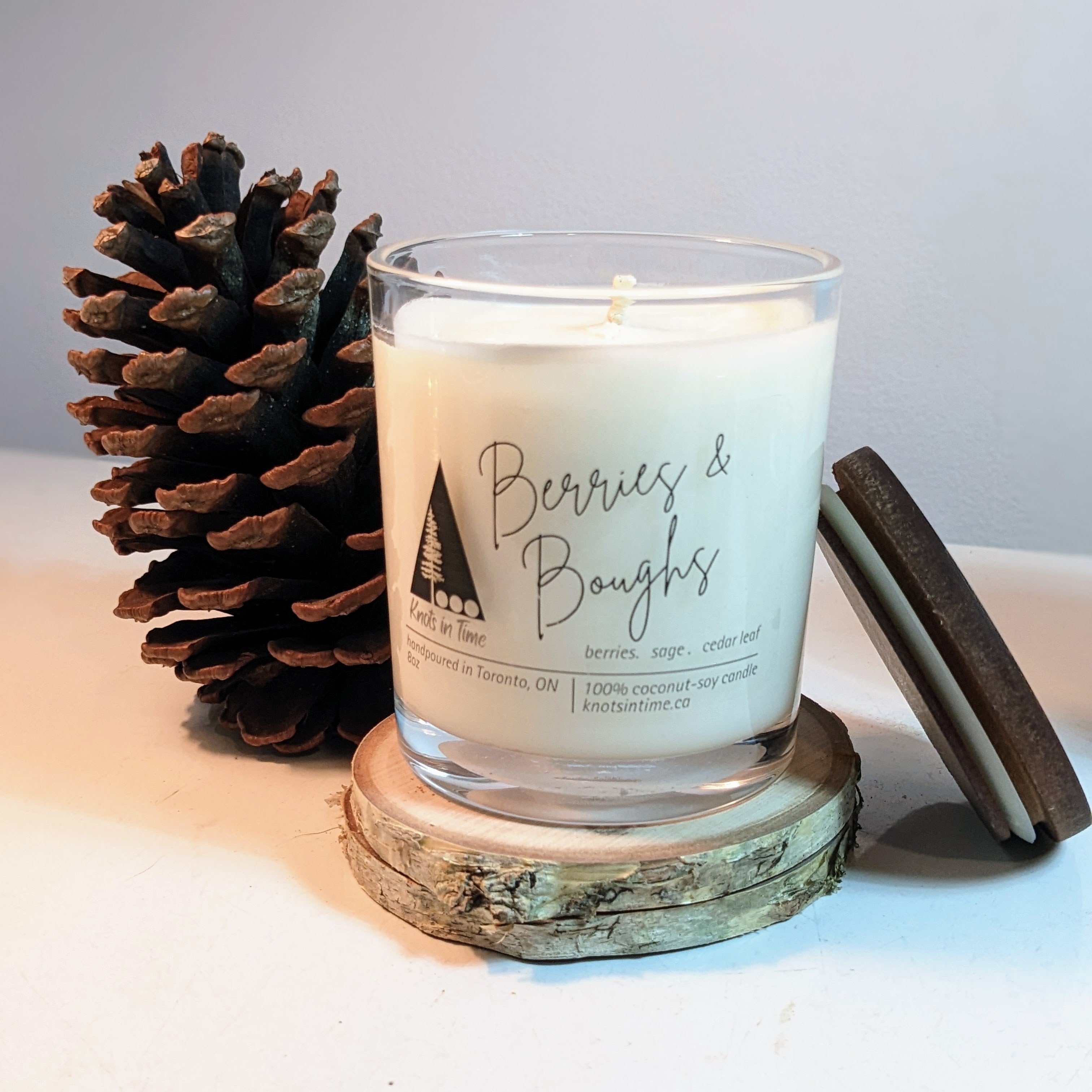 Candle with 'Berries & Boughs' branding next to a pine cone on a light surface.