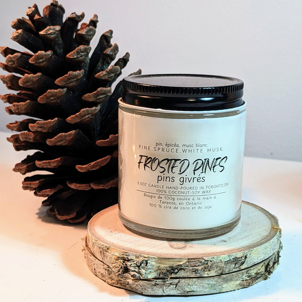 Candle labeled 'Frosted Pines' with a pine cone on a wooden surface