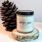 Candle labeled 'Frosted Pines' with a pine cone on a wooden surface