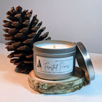 Frosted Pines candle in a tin with a pine cone on a wooden coaster.