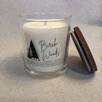 Candle in a glass jar labeled 'Birch Woods' with a wooden lid on a textured surface.