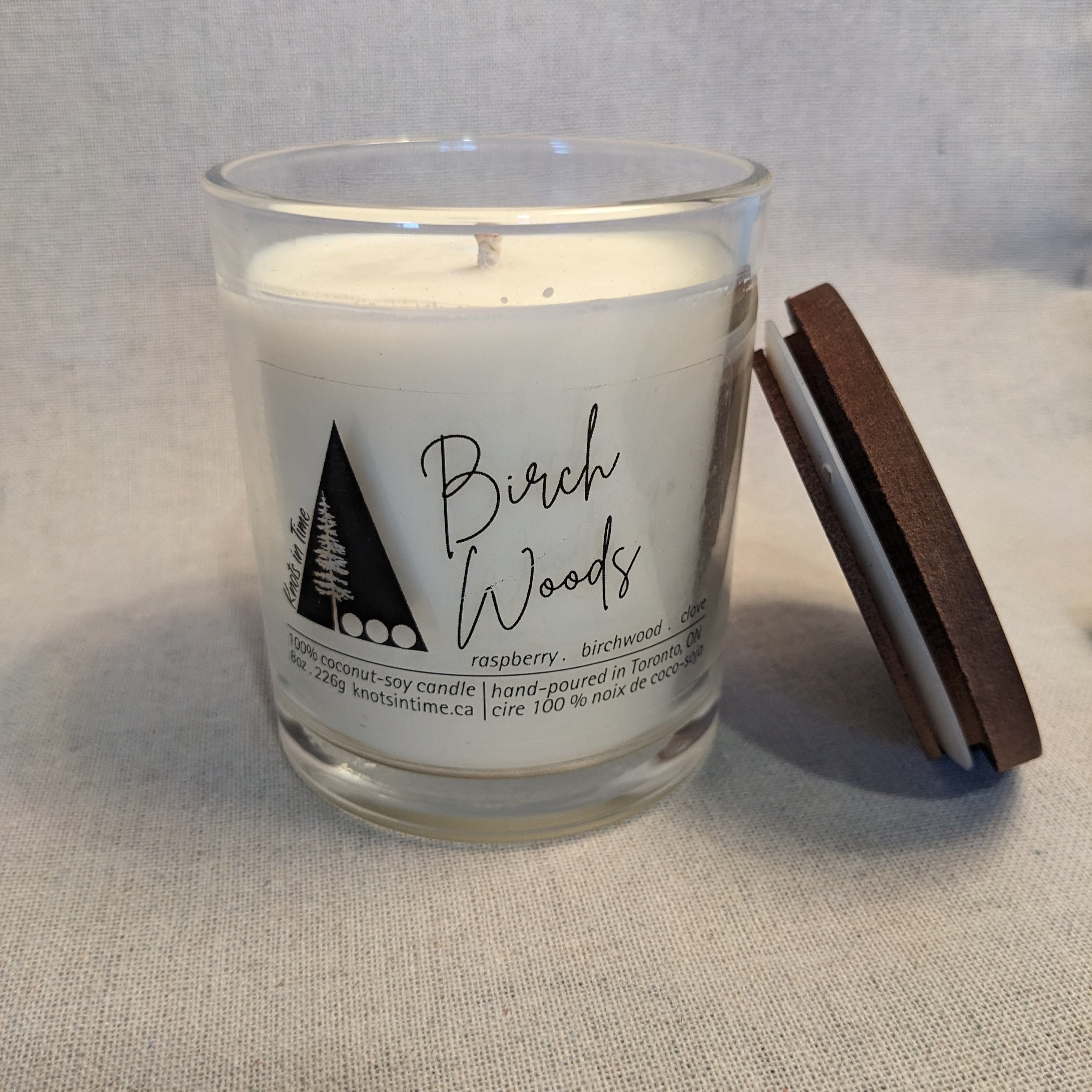 Candle in a glass jar labeled 'Birch Woods' with a wooden lid on a textured surface.