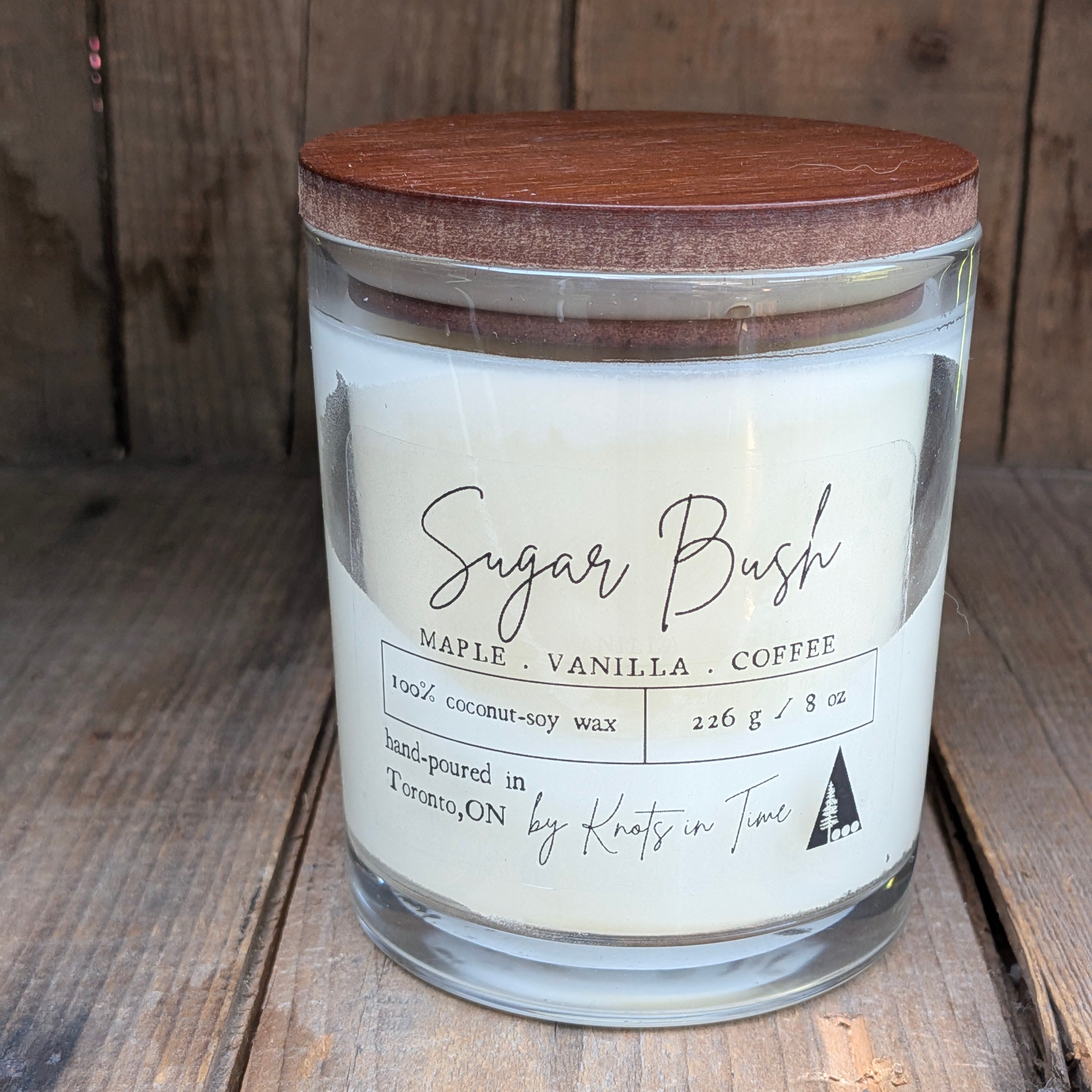 A hand-poured soy wax candle in a clear glass jar with a wooden lid, labeled 'Sugar Bush' by Kats in Time, set against a rustic wooden background.