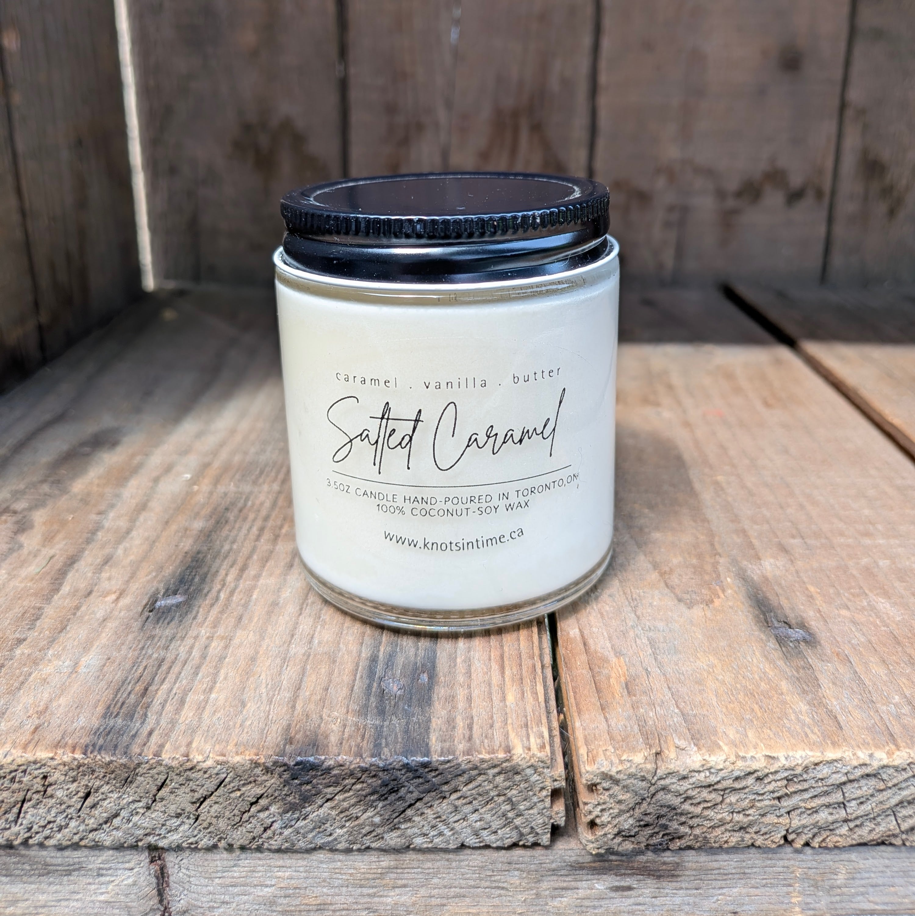 A jar candle labeled 'Salted Caramel' by Knot in Time, sitting on rustic wooden planks. The candle has a white wax fill, a clear glass jar, and a black screw-on lid.