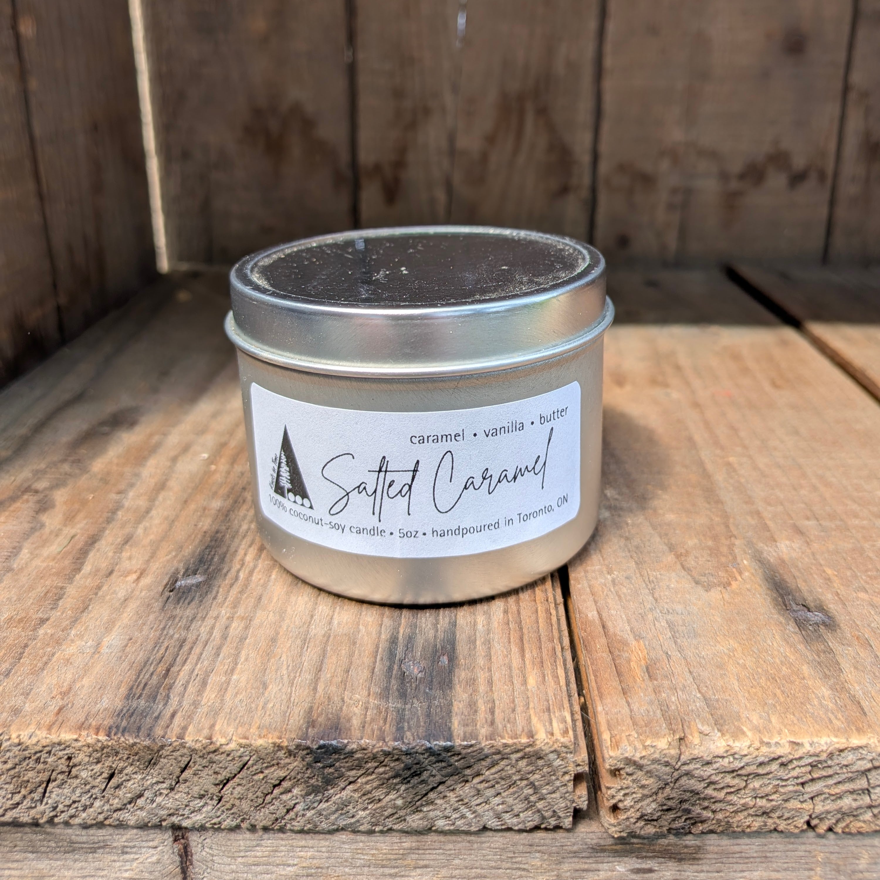 A silver metal tin candle labeled 'Salted Caramel' sits on rustic wooden boards. The label also reads 'caramel • vanilla • butter' and 'handpoured in Toronto, ON'.