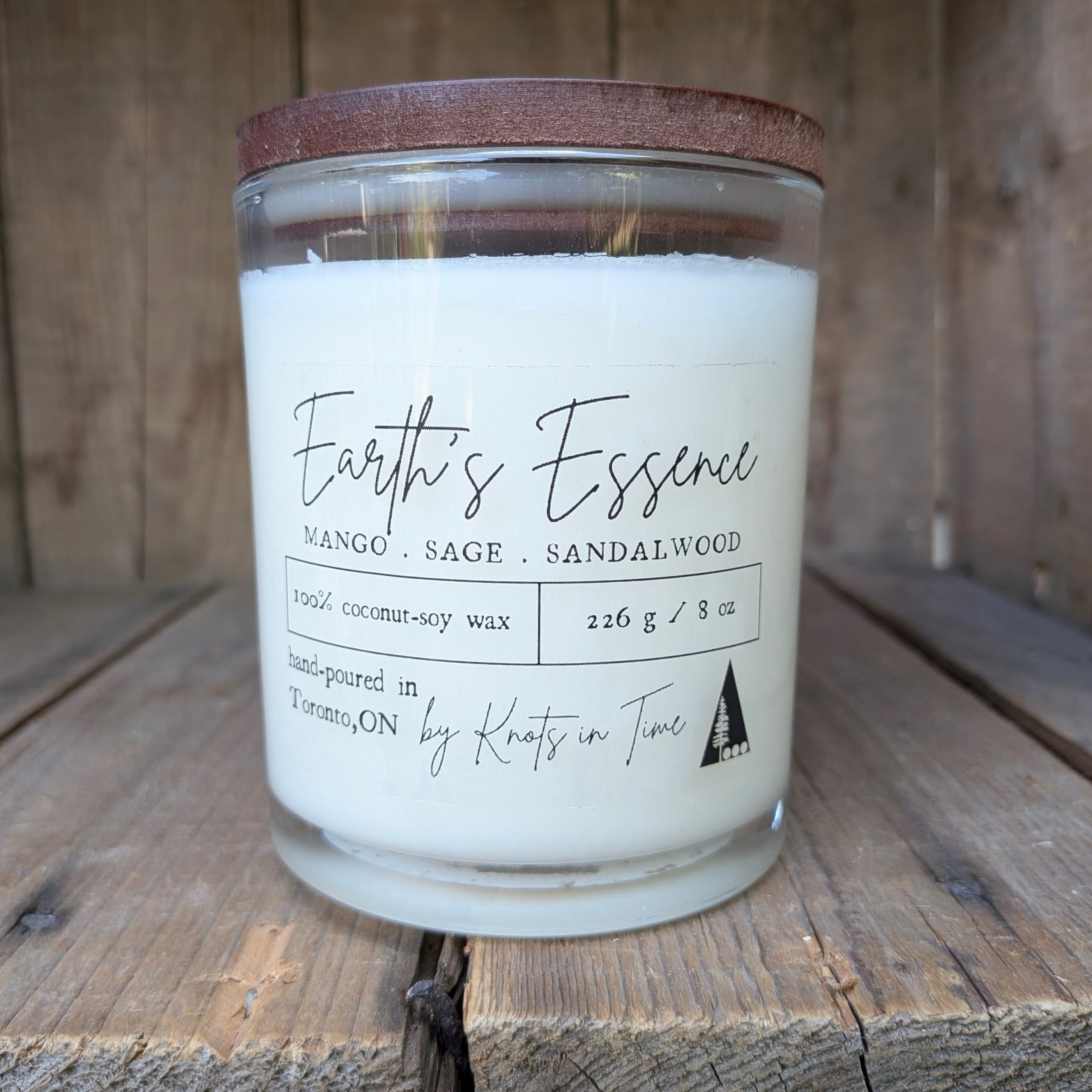 A glass jar candle with white wax and a brown wooden lid, labeled 'Earth's Essence' by Knots in Time, set on a rustic wooden surface against a wooden plank background.