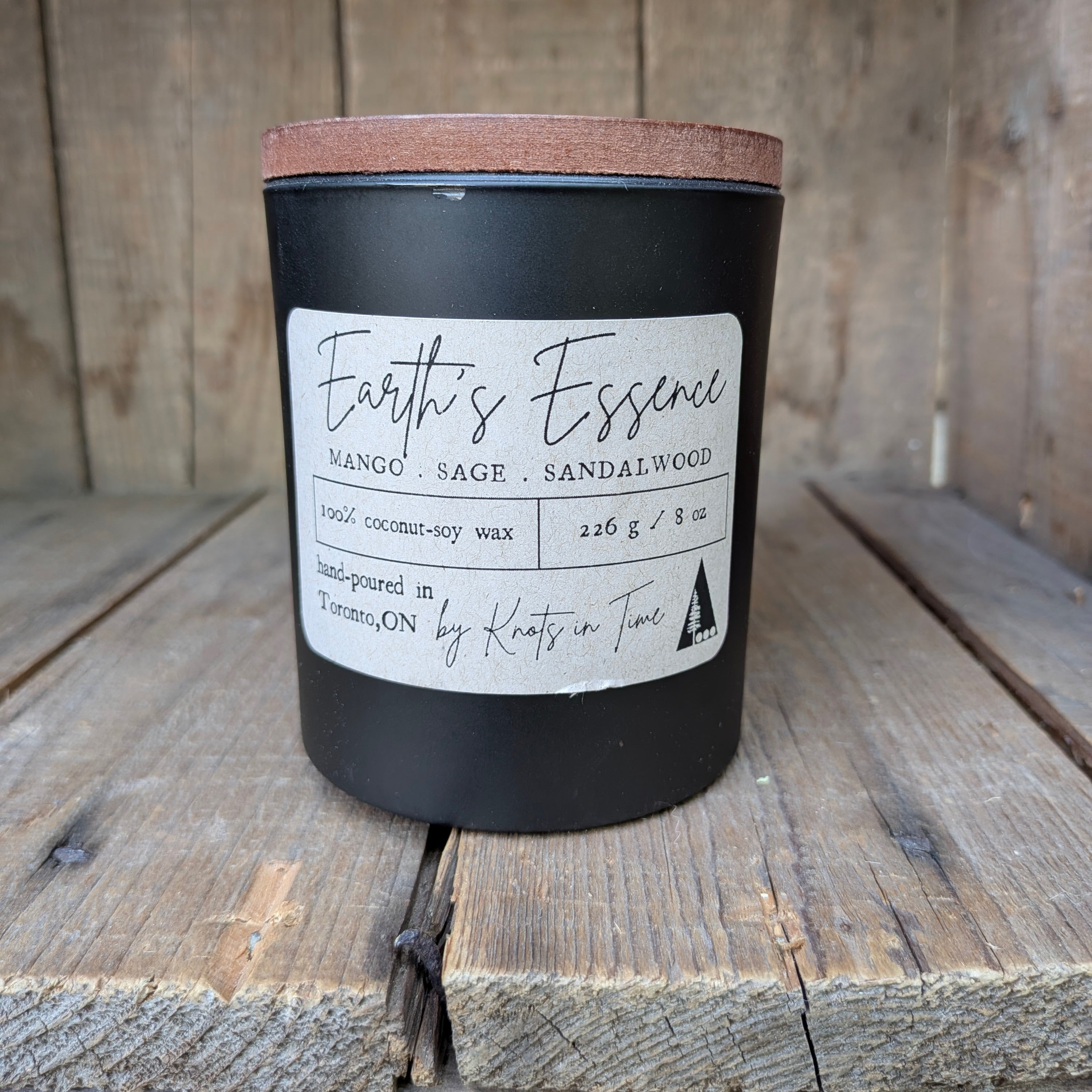 A matte black candle jar with a natural wood lid sits on rustic wooden planks. The front label reads 'Earth's Essence' with scent notes of Mango, Sage, and Sandalwood, and indicates it is hand-poured by Knits in Time.