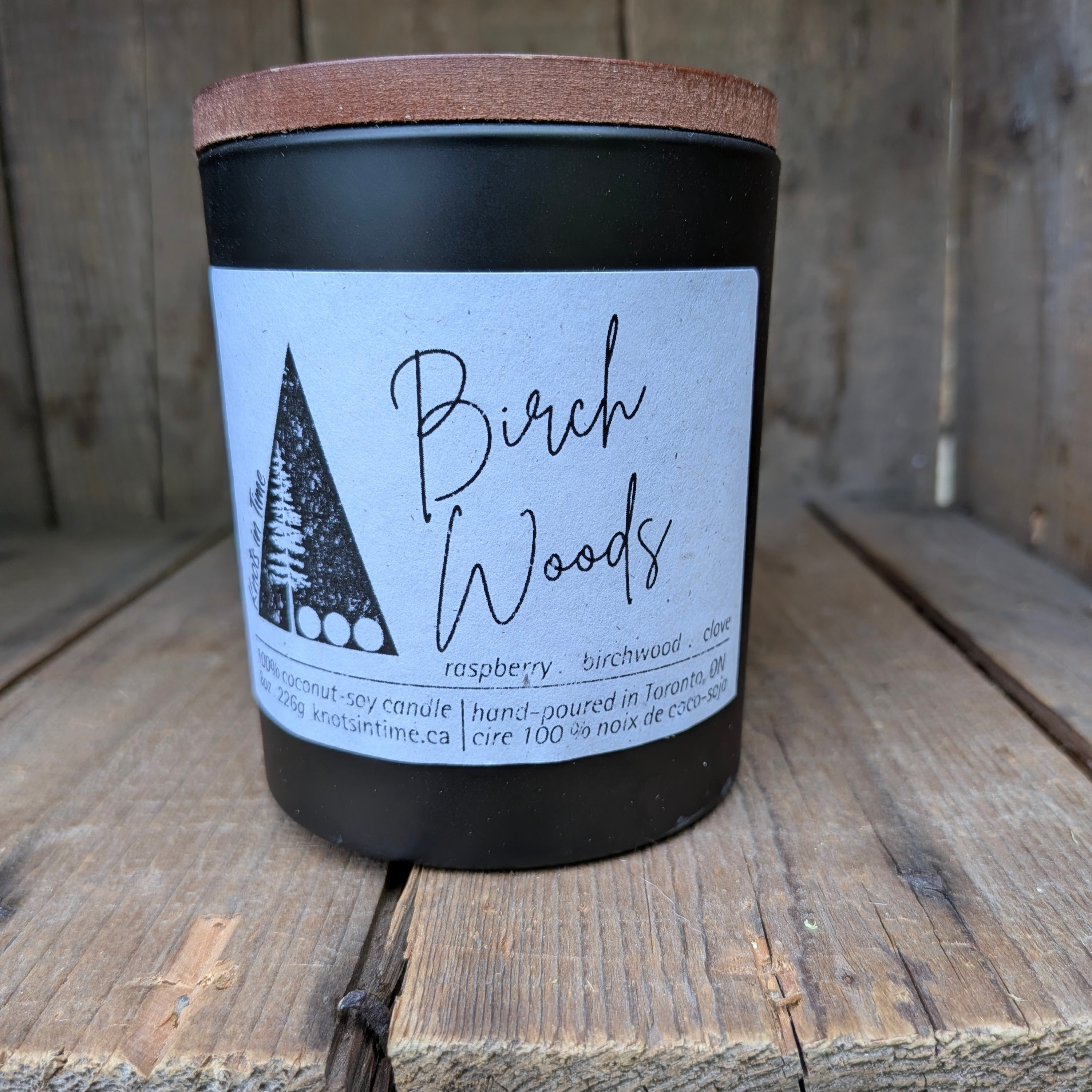 A black jar candle with a light brown wooden lid and a white label reading 'Birch Woods' and 'raspberry, birchwood, clove,' photographed on rustic wooden boards.