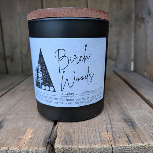A black jar candle with a light brown wooden lid and a white label reading 'Birch Woods' and 'raspberry, birchwood, clove,' photographed on rustic wooden boards.