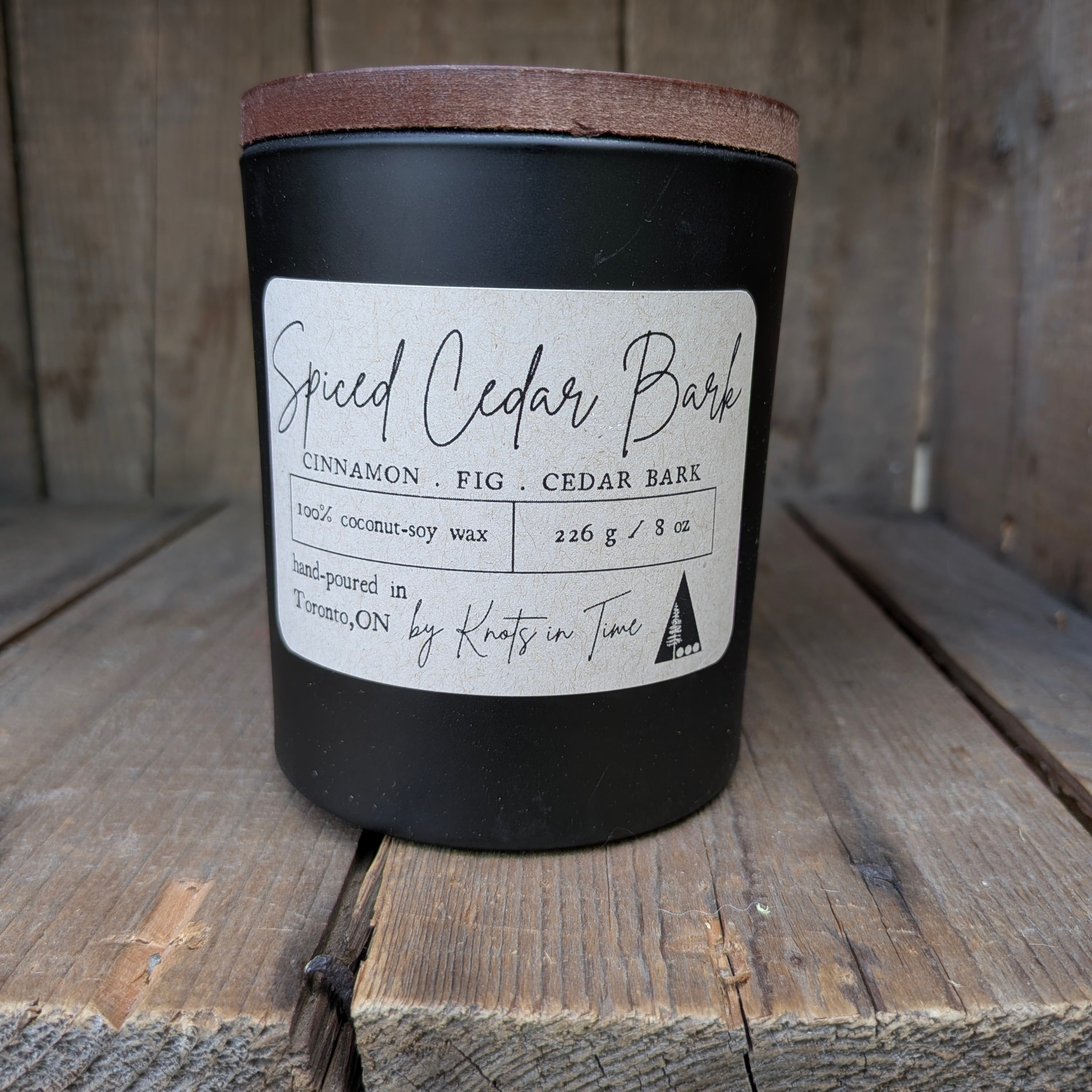 A black jar candle with a brown wood lid sits on rustic wooden planks. The front has a label reading 'Spiced Cedar Bark' with scent notes including cinnamon, fig, and cedar bark, and the brand name Knots in Time at the bottom.