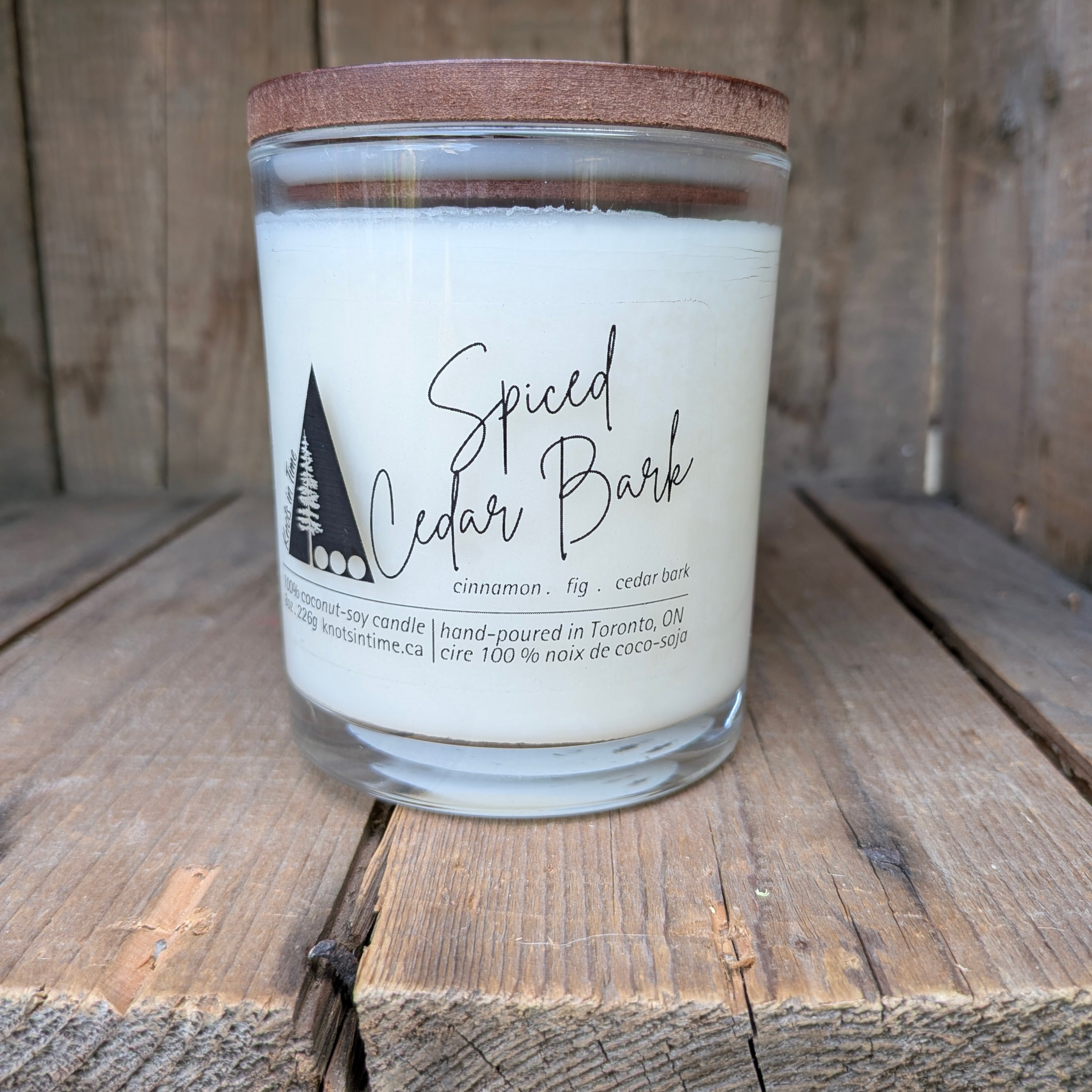 A jar candle labeled 'Spiced Cedar Bark' sits on a rustic wooden surface against a wooden plank background. The candle has a white wax fill, a clear glass jar, and a brown wood style lid.