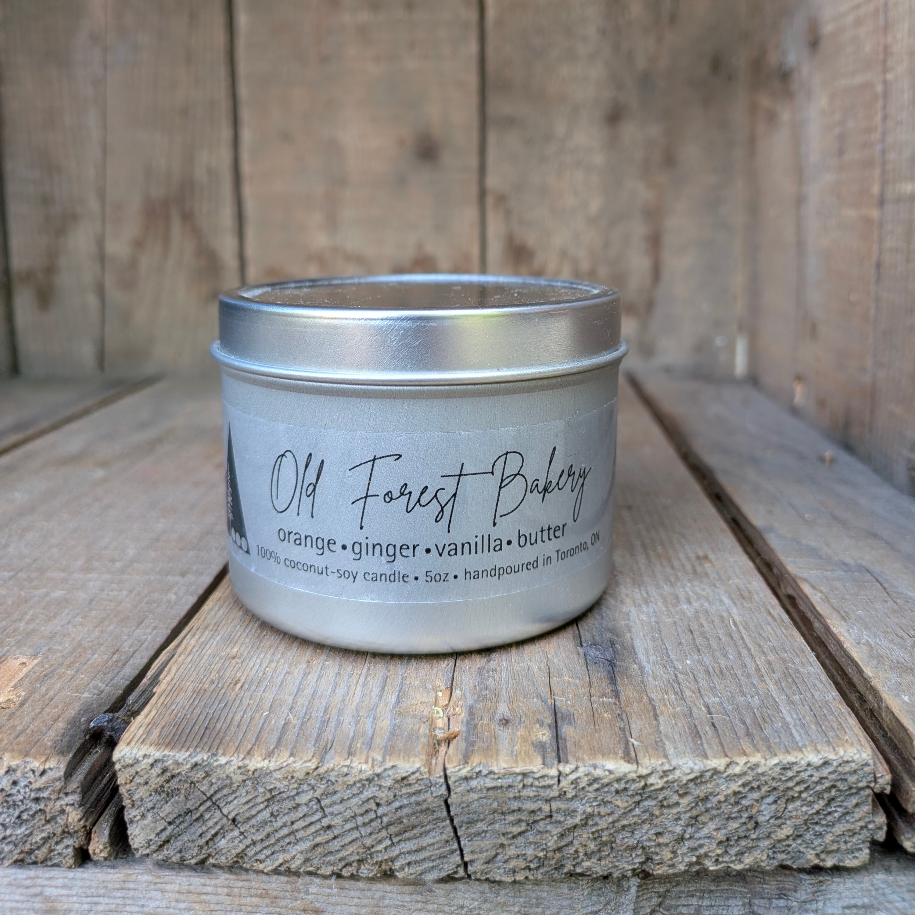 A silver metal candle tin labeled 'Old Forest Bakery' with scent text 'orange • ginger • vanilla • butter,' placed on rustic wooden boards against a wooden plank background.