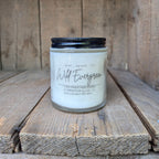 A 3.5oz hand-poured soy wax candle in a clear glass jar with a black metal lid, labeled 'Wild Evergreen' set on rustic wooden planks.