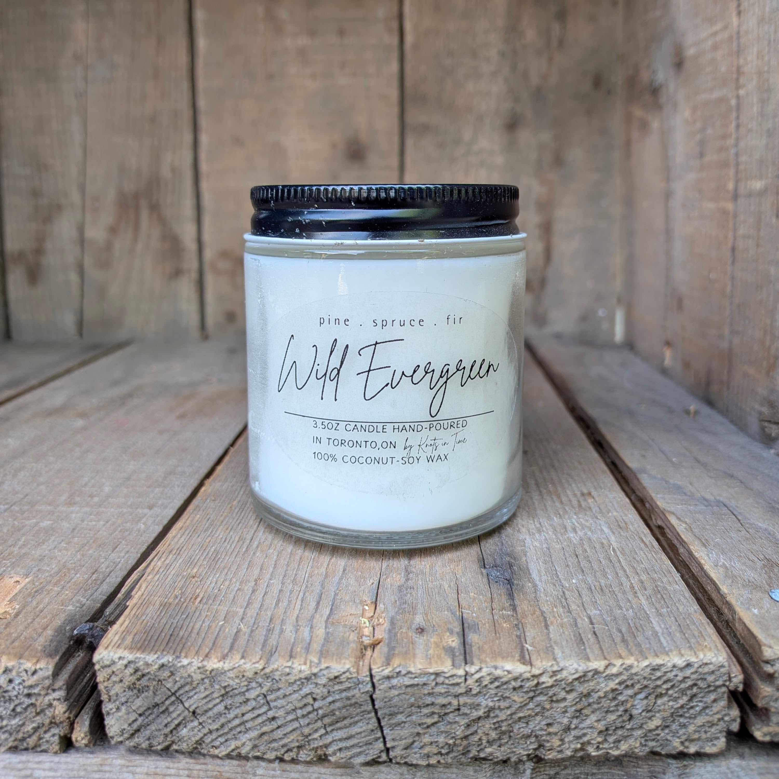 A 3.5oz hand-poured soy wax candle in a clear glass jar with a black metal lid, labeled 'Wild Evergreen' set on rustic wooden planks.