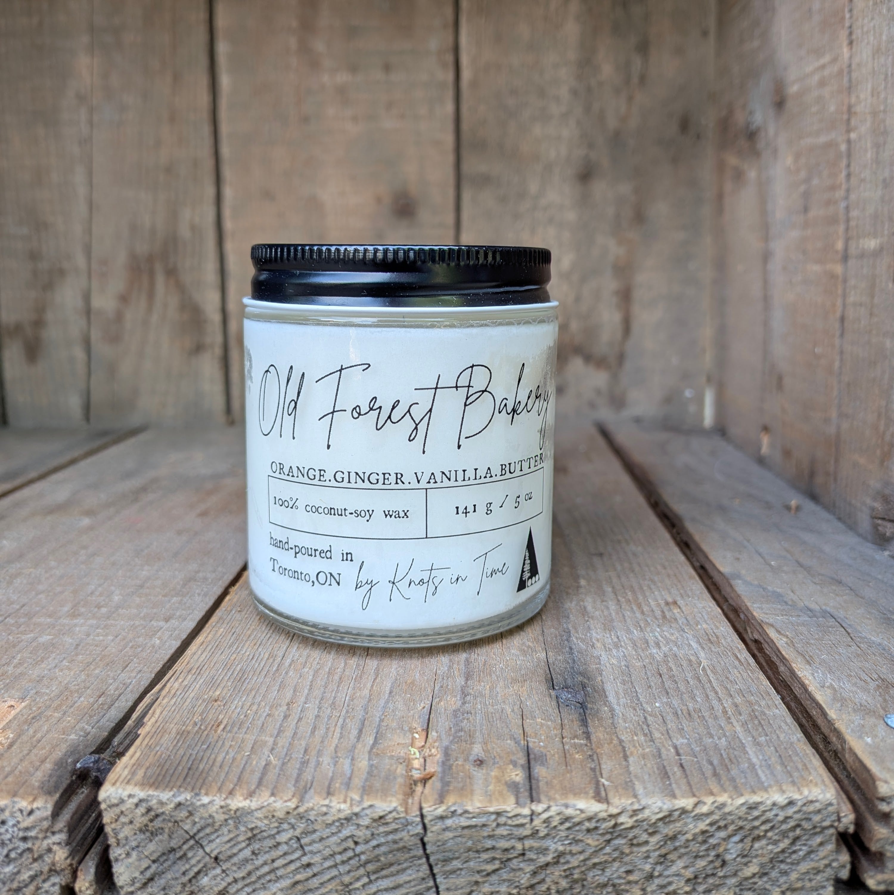 A jar of coconut soy wax candle labeled Old Forest Bakery, featuring a white label with black text and a black metal lid, set on rustic wooden planks.
