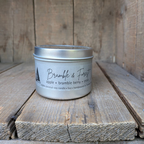 A silver metal tin candle labeled 'Bramble & Forest' with scent notes 'apple • bramble berry • cedar,' placed on rustic wooden boards against a wood plank background.